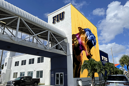 Image of the outside of the building with a large panther mural painted on a large wall and an FIU sign above a skybridge. The door has Mana on one side and FIU on the other in raised letters.